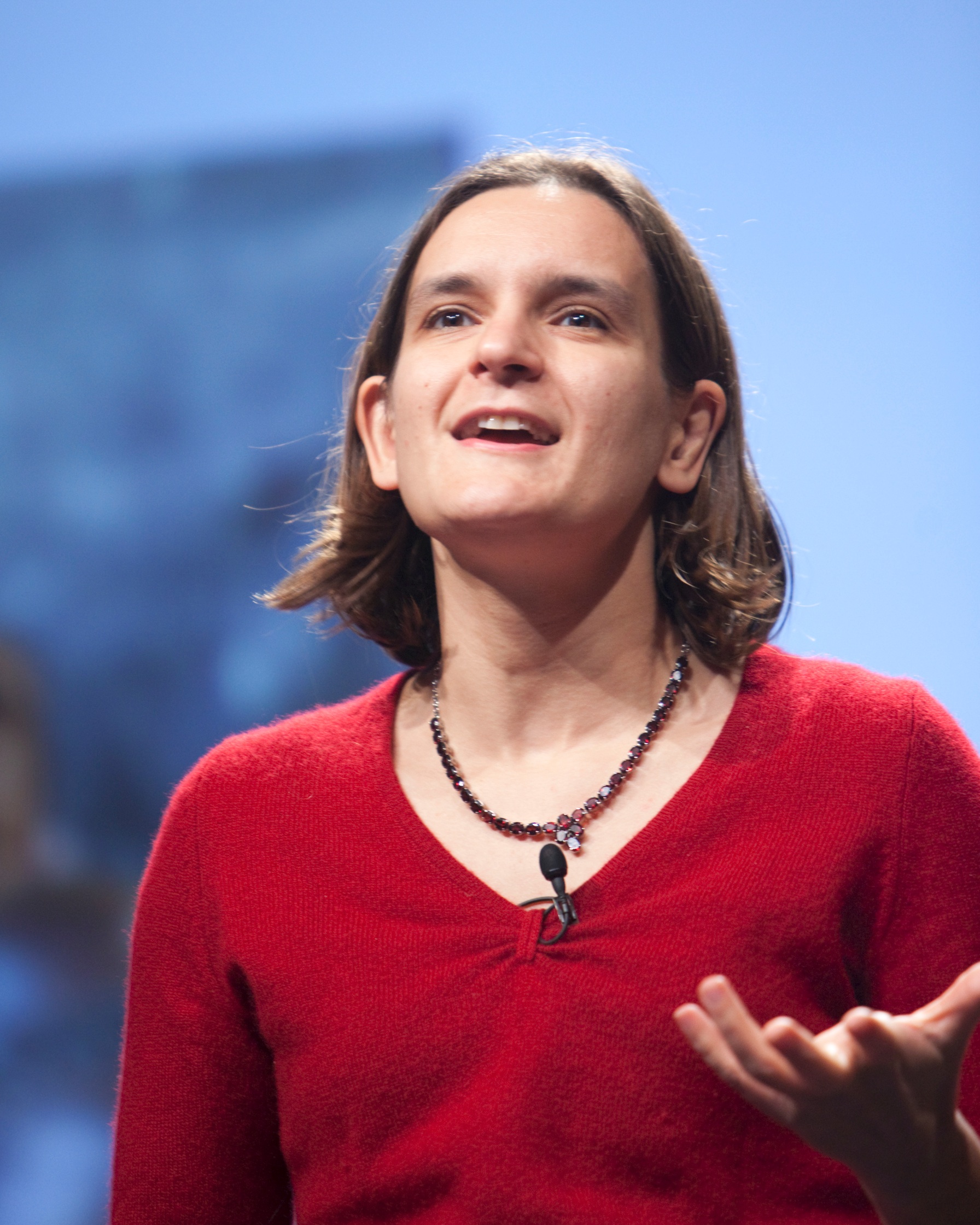 Esther Duflo speaking on stage, wearing a red sweater and necklace, with a microphone clipped to her shirt, expressing herself passionately during a talk.