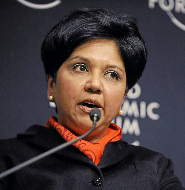 Indra Nooyi speaking at the World Economic Forum in Davos, 2010, wearing a black coat and orange scarf, addressing a global audience with a microphone.