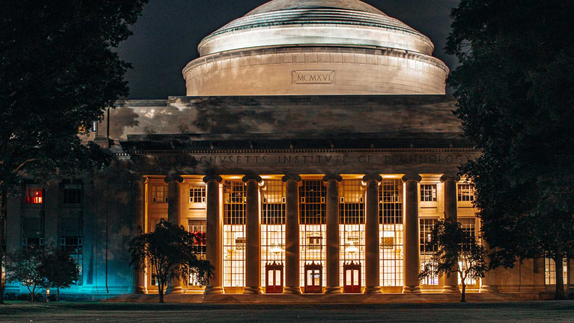 Massachusetts Institute of Technology (MIT) building lit up at night, with iconic dome and classical columns, framed by dark trees.