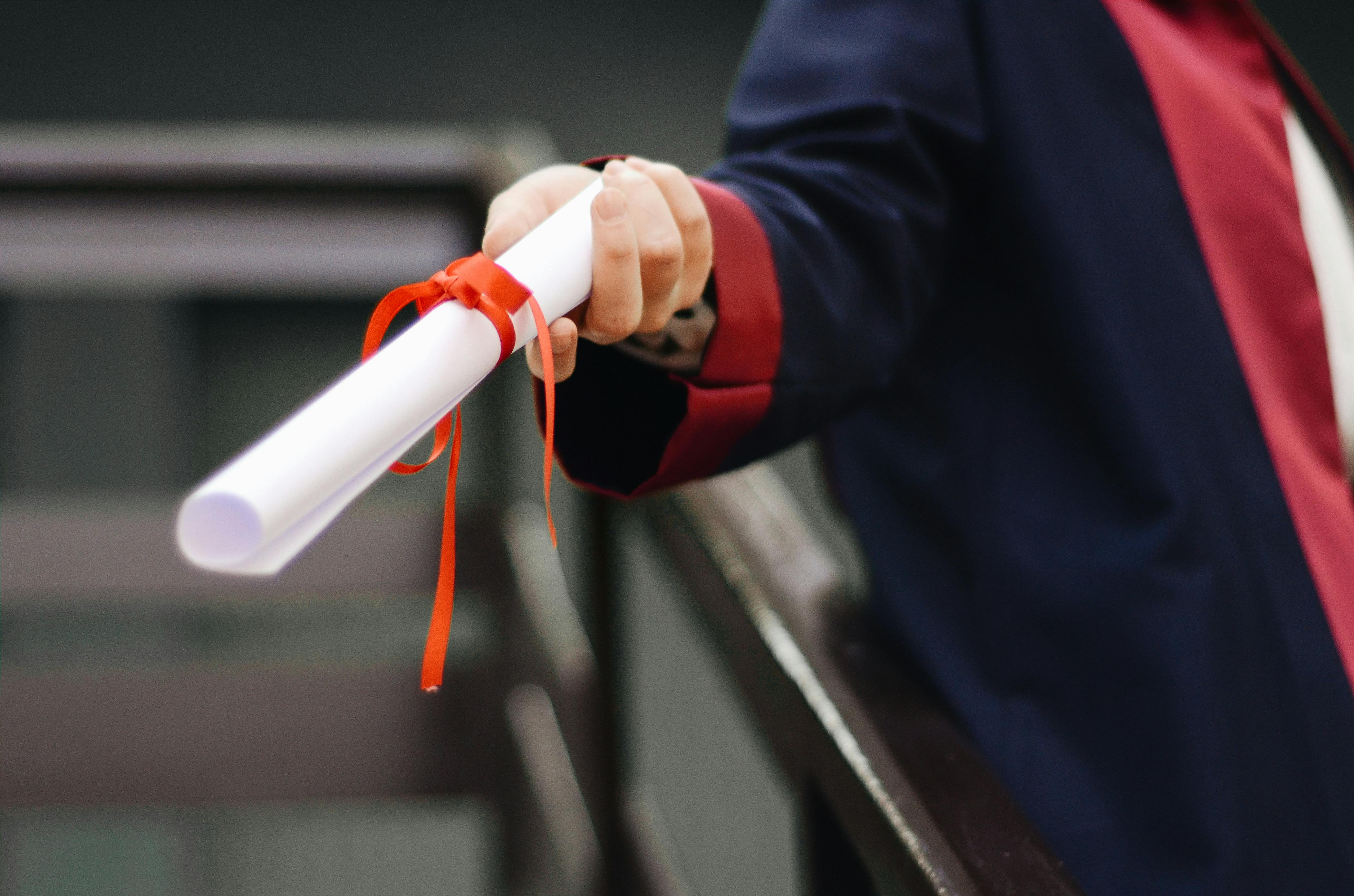 A close-up of a person in a graduation robe handing over a rolled-up diploma tied with a red ribbon, symbolizing achievement, opportunity, and transition.